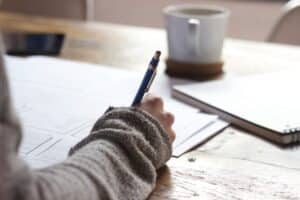 Person writing on paper at a wooden table with a notebook and a coffee cup nearby.